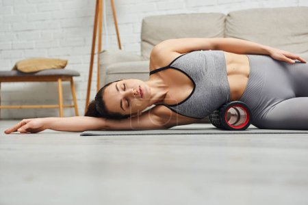 Young brunette and relaxed woman with closed eyes wearing sportswear while using roller massager on body and lying on fitness mat at home, focus on self-care and well-being concept