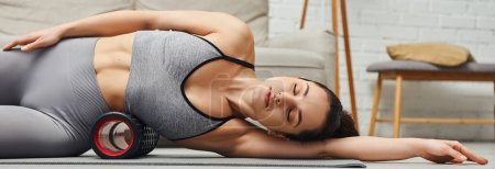 Young and relaxed brunette woman in sportswear massaging body with roller massager and lying on fitness mat at home in living room, focus on self-care and well-being concept, banner 