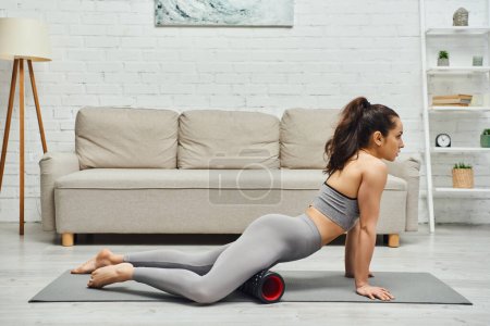 Widok z boku boso i brunetka młoda kobieta w odzieży sportowej masażu nóg z nowoczesnym rolki masażera na macie fitness w domu, utrzymanie zdrowego układu limfatycznego koncepcji, myofascial uwolnienie