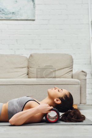 Téléchargez les photos : Vue latérale de la jeune femme brune en vêtements de sport massant le cou avec un masseur à rouleaux moderne sur tapis de fitness dans le salon à la maison, sentiment de tranquillité et promouvoir le concept de relaxation - en image libre de droit