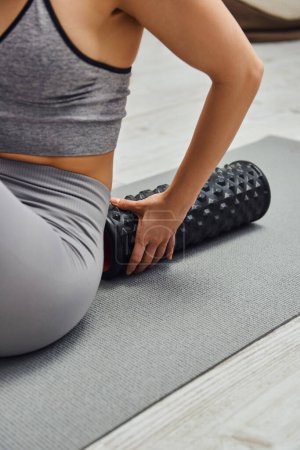Ausgeschnittene Ansicht einer verschwommenen jungen Frau in Sportbekleidung, die einen modernen Rollmassager berührt und zu Hause auf einer Fitnessmatte sitzt, Sinn für Ruhe und Entspannungskonzept fördert, myofasziale Auslösung