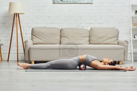 Widok z boku młoda brunetka kobieta w stroju aktywnym relaks leżąc na nowoczesne rolki masażera i maty fitness w salonie w domu, promowanie przepływu limfy i wellness w domu koncepcja