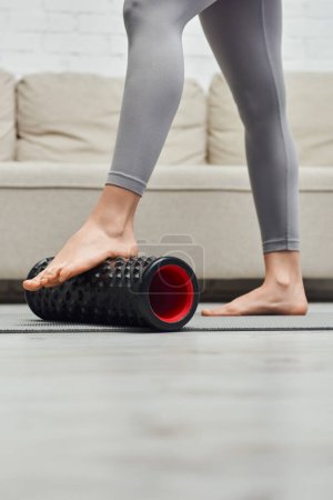 Cropped view of barefoot woman in sportswear massaging feet with modern roller massager on fitness mat near couch in living room, promoting lymph flow and wellness at home concept, tension relief