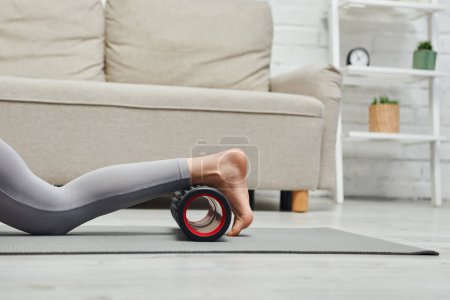 Cropped view of barefoot woman in activewear massaging legs with roller massager on fitness mat near couch in living room at home, promoting lymph flow and wellness at home concept, tension relief