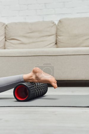 Vista recortada de una mujer descalza que masajea la pierna con un masajeador de rodillos moderno mientras está acostada en una colchoneta de fitness cerca de la sala de estar en casa, promoviendo el flujo linfático y el bienestar en el hogar, alivio de la tensión