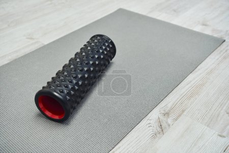 Masajeador de rodillo negro moderno acostado en la colchoneta de fitness en el suelo en casa, promoviendo el flujo linfático y el bienestar en el hogar, la rutina de belleza y bienestar, la salud y la relajación 