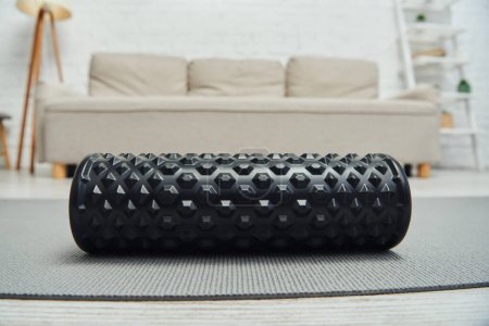 Close up view of black roller massager lying on fitness mat near blurred couch in living room at home, promoting lymph flow and wellness at home concept