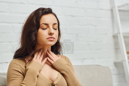 Junge brünette Frau im Pullover überprüft Schilddrüse am Hals, während sie mit geschlossenen Augen auf der Couch im modernen Wohnzimmer zu Hause sitzt, Fokus auf Selbstversorgung und Wohlfühlkonzept, Spannungsabbau
