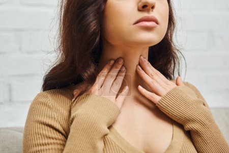 Photo for Cropped view of young brunette woman in brown jumper checking thyroid gland on neck while sitting on blurred couch in living room at home, focus on self-care and well-being concept - Royalty Free Image