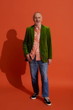 volle Länge des älteren männlichen Modells in grünem Velours-Blazer posiert mit Daumen in der Tasche einer blauen Jeans auf rot-orangefarbenem Hintergrund, lächelndes Gesicht, persönlicher Stil, positives Alterskonzept