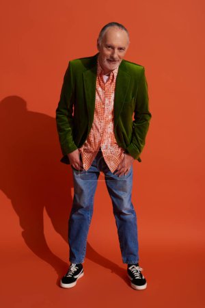 full length of positive elderly man in shirt and green velour blazer holding thumbs in pockets of blue jeans, leaning forward and looking at camera on red orange background, stylish aging concept
