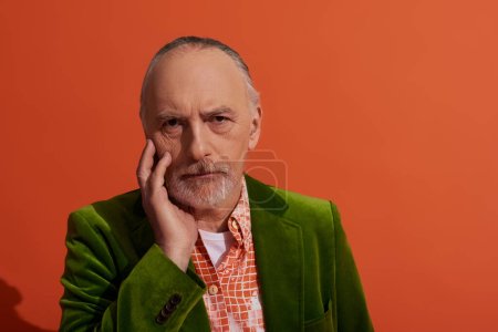 mode et âge, portrait de cheveux gris, barbu, homme réfléchi touchant le visage et regardant la caméra sur fond rouge orange, modèle masculin senior, concept de population vieillissante
