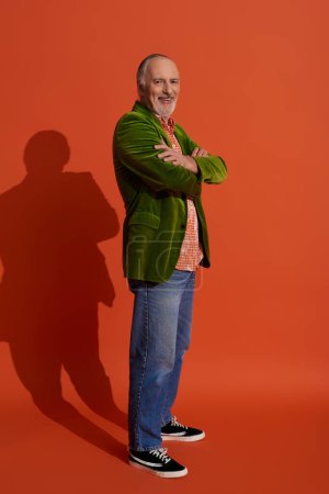 full length of handsome and bearded senior man standing with folded arms and smiling at camera on red orange background with shadow, green velour blazer, blue denim jeans, stylish aging concept