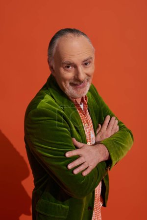 Photo for Optimistic and charismatic bearded senior man standing with folded arms and smiling at camera on red orange background, fashion look, personal style, green velour blazer, happy aging concept - Royalty Free Image