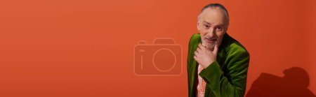 curious and joyful senior man with grey hair and beard holding hand near chin and looking at camera on red orange background, older model, green velour blazer, positive and stylish aging, banner