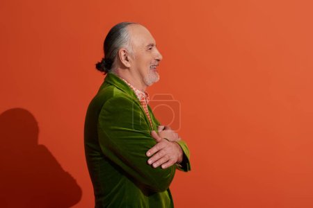 Foto de Vista lateral del modelo masculino senior, hombre optimista posando con los brazos cruzados y sonriendo sobre fondo rojo anaranjado, chaqueta de terciopelo verde, pelo gris y barba, concepto de envejecimiento positivo y de moda - Imagen libre de derechos
