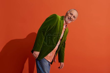 charismatic and carefree senior model posing on red orange background with shadow and smiling at camera, trendy casual clothes, green velour blazer, personal style and positive aging concept