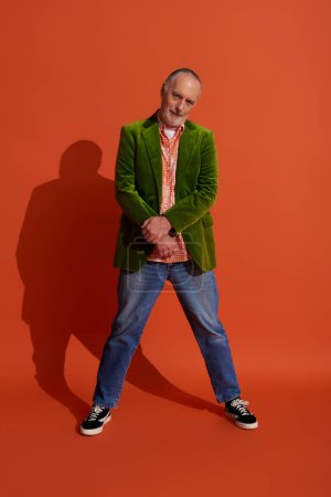 volle Länge des coolen und selbstbewussten älteren Mannes in stylischer Freizeitkleidung stehend und in die Kamera blickend auf rot-orangefarbenem Hintergrund mit Schatten, grüner Velours-Blazer, blaue Jeans, Modelook