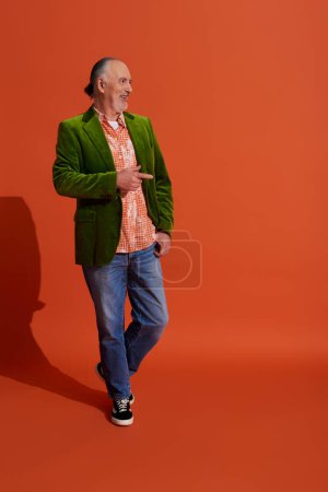 volle Länge des coolen und fröhlichen älteren Mannes in grünem Velours-Blazer, der mit der Hand in der Tasche einer blauen Jeans steht, wegschaut und mit dem Finger auf rotorangefarbenen Hintergrund mit Schatten zeigt