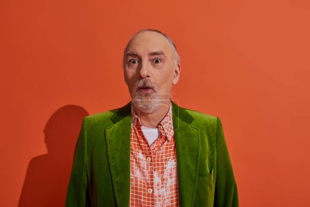 surprised and impressed senior man with bulging eyes looking at camera on red orange background, grey hair and beard, green velour blazer and trendy shirt, personal style and positive aging concept