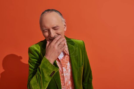 happy aging, overjoyed senior man covering mouth with hand and laughing with closed eyes on red orange background, green velour blazer, trendy shirt, fashion and age concept