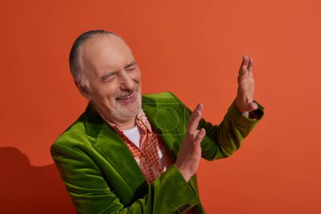 aufgeregter grauhaariger und bärtiger älterer Herr im grünen Velours-Blazer mit Stop-Geste und Lachen mit geschlossenen Augen auf rot-orangefarbenem Hintergrund, persönlicher Stil, modisches Alterskonzept