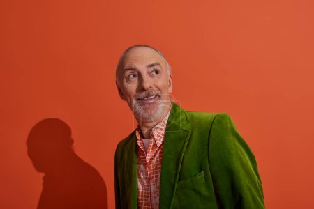 joyful and surprised senior man with grey hair and beard smiling and looking away on red orange background, fashionable casual clothes, green velour blazer, trendy shirt, positive aging concept