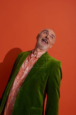 glücklich alterndes, trendiges Seniormodel, überglücklicher Mann mit grauen Haaren und Bart, lachend und wegschauend auf rot-orangefarbenem Hintergrund, grüner Velours-Blazer, trendiges Hemd, persönlicher Stil