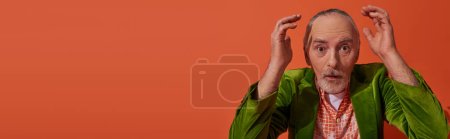 dazed senior man with grey hair, beard and bulging eyes holding hands near head and looking at camera on red orange background, trendy green velour blazer, personal style, banner with copy space