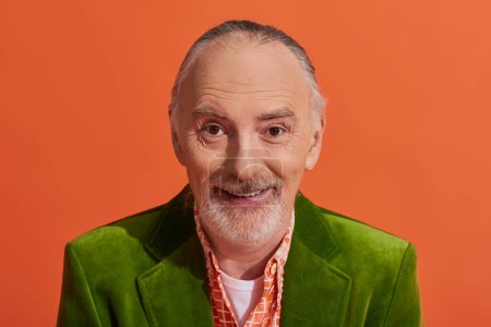 portrait de charismatique modèle senior masculin avec cheveux gris, barbe et sourire radieux en regardant la caméra sur fond orange vif, veste en velours vert, vêtements décontractés à la mode, vieillissement positif