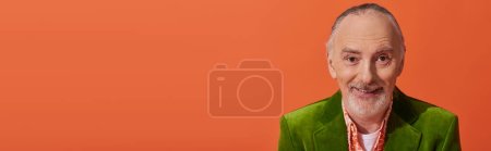 portrait of trendy and cheerful senior model with grey hair and beard, wearing green velour blazer and smiling at camera on vibrant orange background, fashion and age concept, banner with copy space
