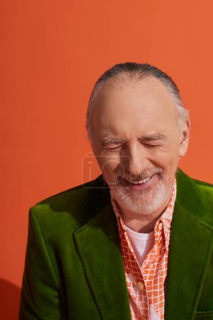 happiness, portrait of overjoyed senior and bearded man in green velour blazer laughing with closed eyes on vibrant orange background, fashion look, positive and stylish aging concept