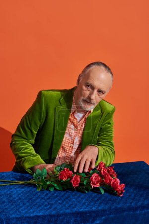 gealterter, nachdenklicher Mann am Tisch sitzend mit blauem Tuch und roten Rosen, während er vor leuchtend orangefarbenem Hintergrund wegschaut, älteres, bärtiges Model im grünen Samtblazer, trendiges alterndes Konzept
