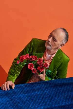 memories, melancholy, bearded senior man in green velvet blazer holding bouquet of red roses while sitting at table with blue velour cloth on vibrant orange background, aging population concept