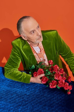 nachdenklicher und modischer älterer Herr in grünem Samtblazer sitzt mit Strauß roter Rosen am Tisch mit blauem Velourtuch und schaut weg auf leuchtend orangefarbenen Hintergrund, stilvolles Alterskonzept