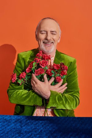 overjoyed senior man in green velvet blazer hugging red roses and smiling with closed eyes near table with blue velour cloth on vibrant orange background, positive aging concept
