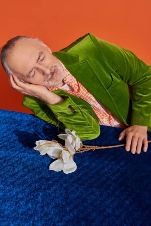 glückliche Erinnerungen, älterer Herr sitzt neben weißer Orchidee auf blauem Velourtisch und lächelt mit geschlossenen Augen auf leuchtend orangefarbenem Hintergrund, grünem Samtblazer, positivem Alterungskonzept, Blickwinkel hoch