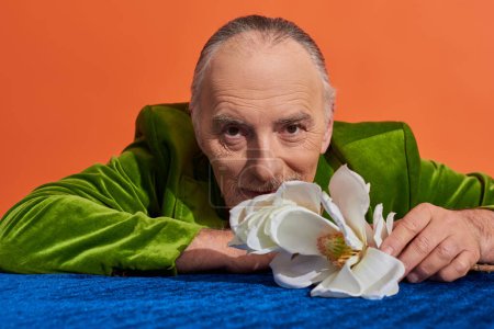 positiver Lebensstil, älterer grauhaariger Mann in grünem Samtblazer lächelt in die Kamera neben weißer Orchideenblume auf blauem Velourtuch auf leuchtend orangefarbenem Hintergrund, glückliches und stilvolles Alterskonzept