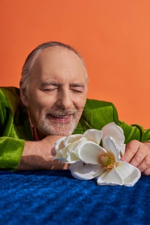 pleasant memories, stylish senior man in green velvet blazer smiling with closed eyes near white orchid flower on blue velour cloth on vibrant orange background, happy lifestyle and aging concept