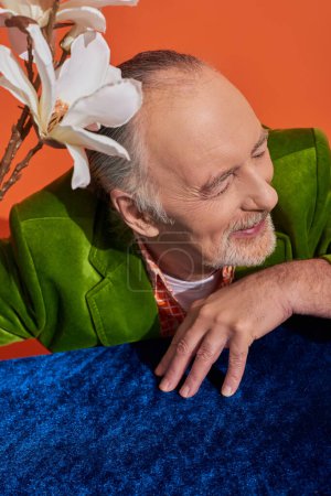 cheerful bearded and grey haired senior man in green velvet blazer sitting with closed eyes near white orchid and table with blue velour cloth on vibrant orange background, happy aging concept