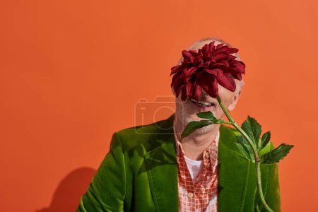 optimistisches und trendiges Seniormodel in grünem Velours-Blazer, der sich hinter roter Pfingstrose versteckt, während er auf leuchtend orangefarbenem Hintergrund sitzt, älterer Mann in grünem Samt-Blazer, glückliches alterndes Konzept