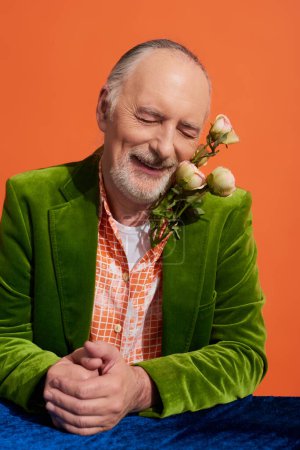 zufriedener älterer Mann mit grauen Haaren, Bart und geschlossenen Augen, der mit frischen Rosen posiert und am Tisch mit blauem Velourtuch auf leuchtend orangefarbenem Hintergrund lächelt, grüner Samtblazer, stylisches Alterskonzept