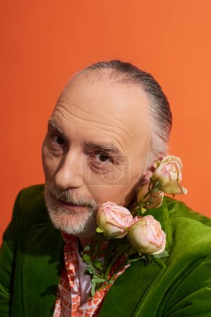 portrait of positive grey haired man with groomed beard and expressive gaze posing with roses and looking at camera on vibrant orange background, green velour jacket, fashionable aging concept
