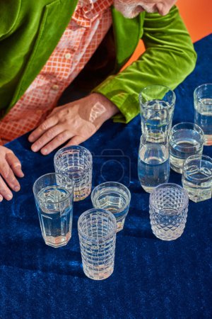cropped view of senior man in trendy and green velvet blazer sitting near crystal glasses with clear water on table with blue velour cloth on orange background, symbolism, life fullness concept