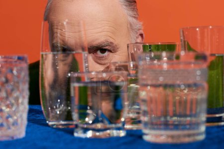 Senior männliches Modell mit ausdrucksstarkem Blick in die Kamera hinter verschwommenen Kristallgläsern mit reinem Wasser auf orangefarbenem Hintergrund, alternde Bevölkerung, Symbolik, Lebensfüllungskonzept