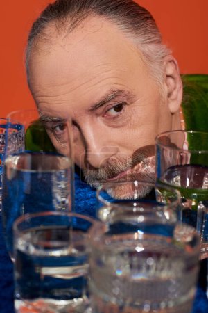 Erinnerungen, älterer und nachdenklicher Mann, der in der Nähe von Gläsern mit klarem Wasser auf einem Tisch mit blauem Velourtuch auf orangefarbenem Hintergrund wegschaut, alternde Bevölkerung, Symbolik, Lebensfüllungskonzept