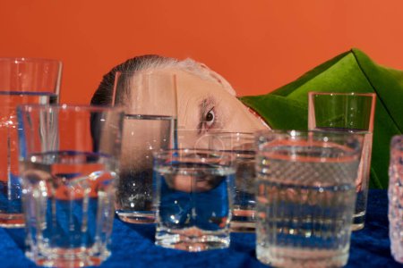 älterer Mann, der sein Gesicht hinter durchsichtigen Gläsern mit klarem Wasser auf einem Tisch mit blauem Velourtuch auf orangefarbenem Hintergrund verdeckt, alternde Bevölkerung, Symbolik, Lebensfüllungskonzept
