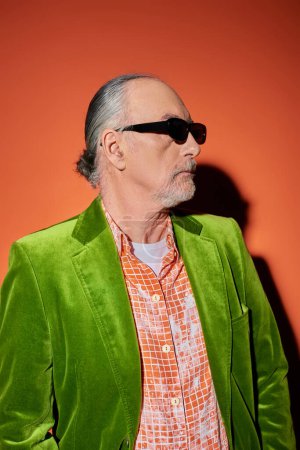 Mode-Look, älteres männliches Model mit grauen Haaren und Bart auf rotem und orangefarbenem Hintergrund mit Schatten, dunkle Sonnenbrille, trendiges Hemd, grüner Velours-Blazer, stilvoller Lebensstil alternder Männer