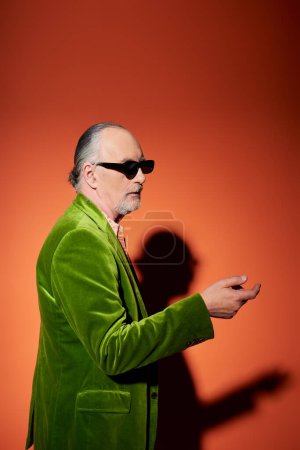 älterer, grauhaariger Mann, der mit der Hand zeigt und vor rotem und orangefarbenem Hintergrund mit Schatten wegsieht, dunkel stylische Sonnenbrille, grüner Velours-Blazer, Individualität, trendiges alterndes Konzept