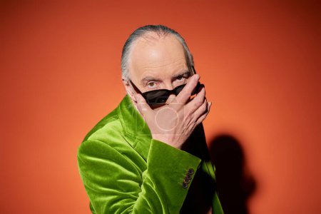 Photo for Elderly and fashionable man in green velour blazer taking off dark stylish sunglasses and looking at camera with expressive gaze on red and orange background, trendy aging concept - Royalty Free Image